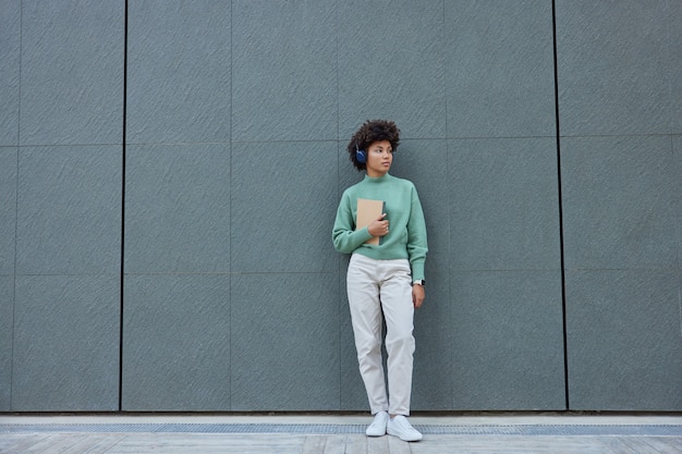 girl dressed in fashionable clothes looks away listens music in headphones holds diary poses against grey wall