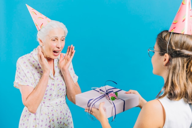 Girl giving birthday gift to surprised grandmother on blue backdrop