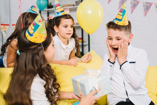 Girl giving wrapped gift box to surprised birthday boy in the party