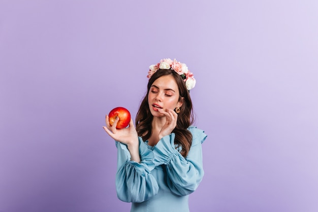 Free photo girl in great mood is looking at delicious red apple. snapshot of woman in blue blouse on isolated wall.