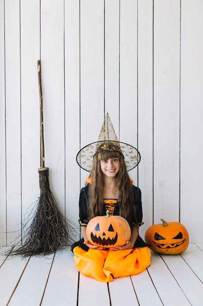 Girl in Halloween costume sitting in studio with pumpkins and broom