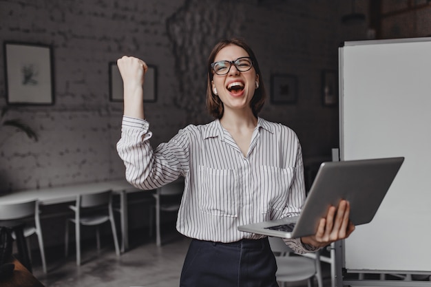 Girl happily screams and makes winning hand gesture, holding laptop and posing in office against background of board.