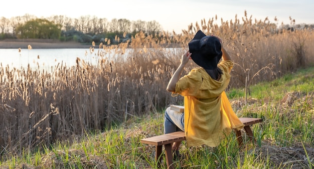 A girl in a hat and in casual style sits on a bench near the lake at sunset.