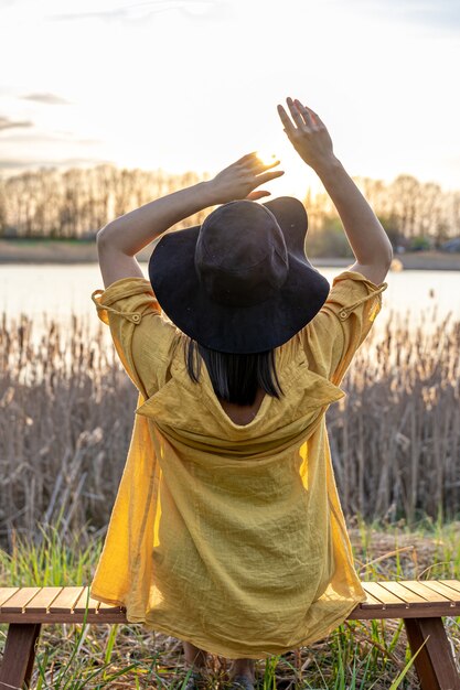 A girl in a hat and in casual style sits on a bench near the lake at sunset