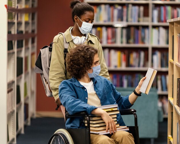 Girl helping her colleague in wheelchair choose his books in the library