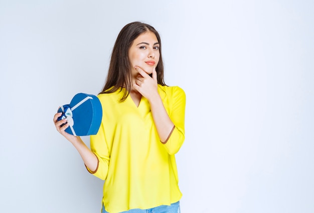 Girl holding a blue heart shape gift box and thinking or hesitating.