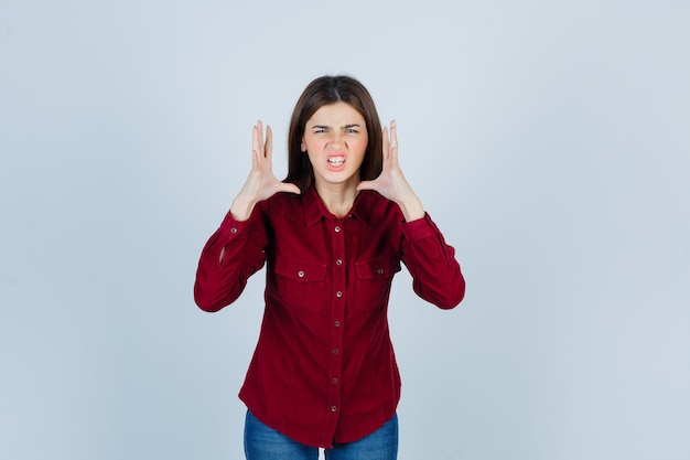 girl holding hands near head in burgundy shirt and looking aggressive.