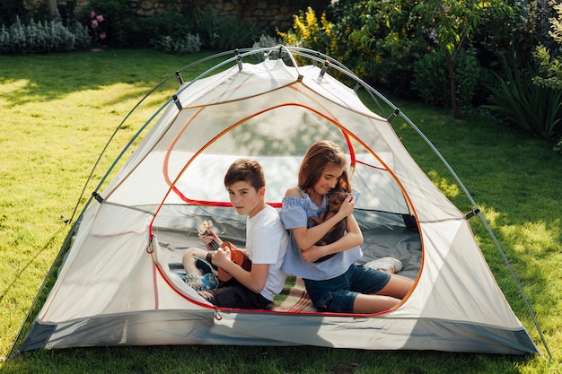 Girl holding little dog sitting with her brother in tent camp in park