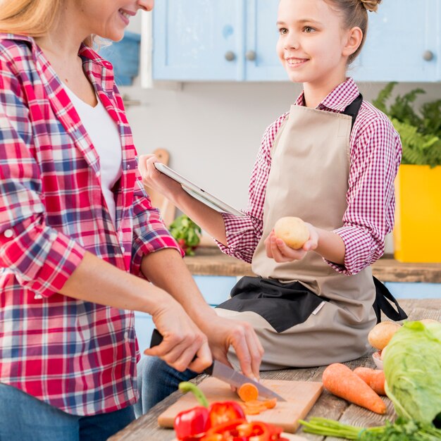 Girl holding potato and digital tablet in hand looking at her mother cutting vegetables with knife