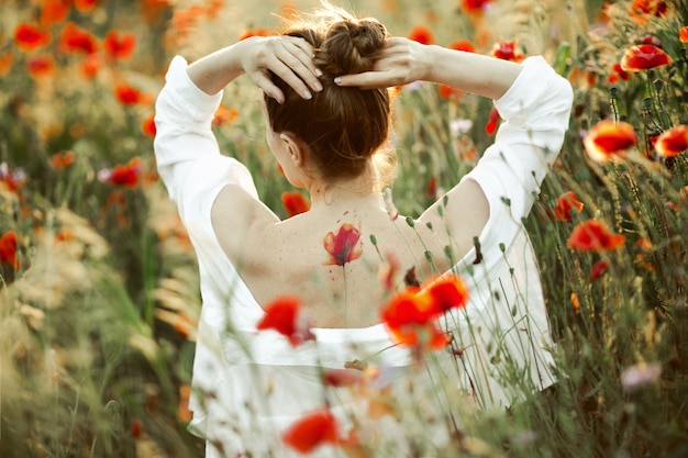 Free Photo girl holds hands for head and stands with naked back with a tattoo flower poppy on it, among the poppies field