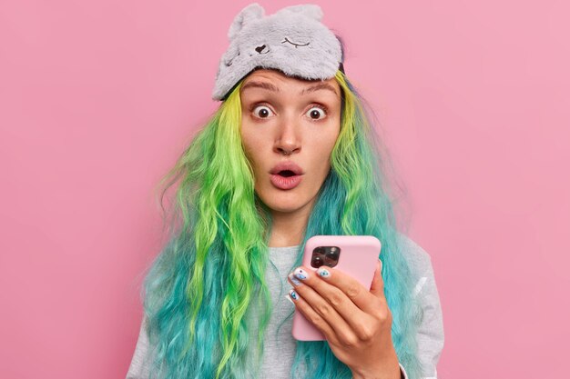  girl holds mobile phone stares bugged eyes at camera cannot believe in shocking news has dyed long hair reads message on cellular has surprised face wears pajama with blindfold