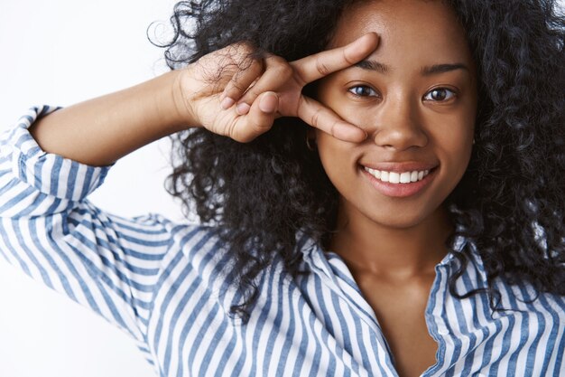 Girl likes anime showing peace sign. Portrait emotive carefree happy dark-skinned young woman curly hairstyle make disco victory gesture near eye feeling awesome joyful, standing white wall