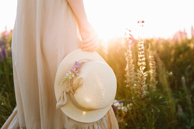 Free photo girl in long dress holding straw hat standing in spring flower field at sunset