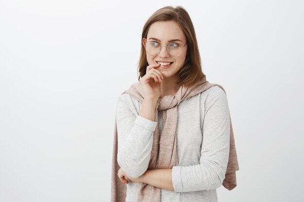 Girl looking at curious guy trying turn attention biting finger and smiling intrigued gazing left with interest and desire having mysterious delightful idea standing over gray wall