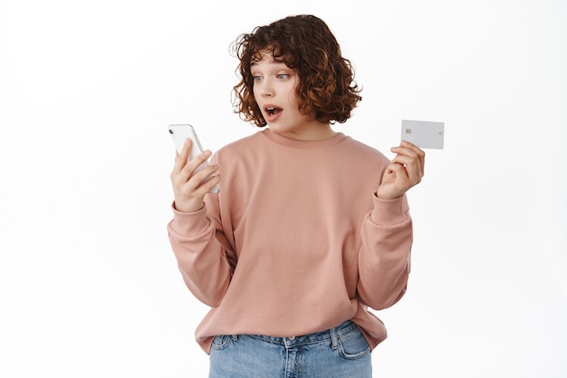 Girl looks surprised at mobile phone screen while shopping online, holding credit card, paying for internet order delivery, spot good price in shop, standing on white