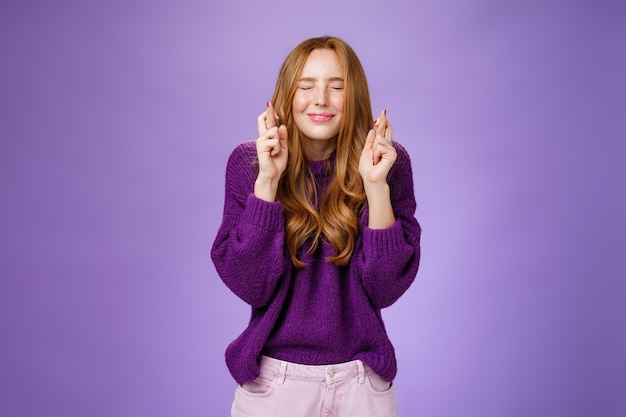 Free photo girl makes wish eager dream come true. atractive hopeful and positive young redhead woman in purple sweater close eyes and smiling as cross fingers for good luck, praying and anticipating miracle.