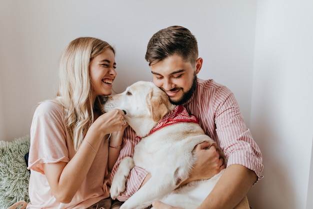 Girl in oversize shirt laughs wholeheartedly and looks at her dark-haired man holding Labrador.