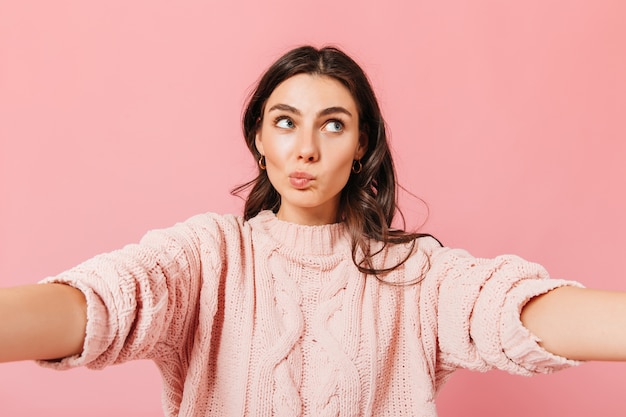 Girl in oversize sweater looks up thoughtfully. Curly brunette takes selfie on pink background and wants hugs.