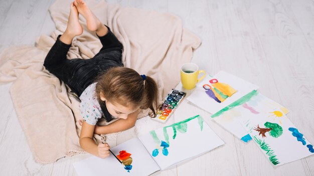 Free Photo girl painting by water colors on paper near draws and lying on floor