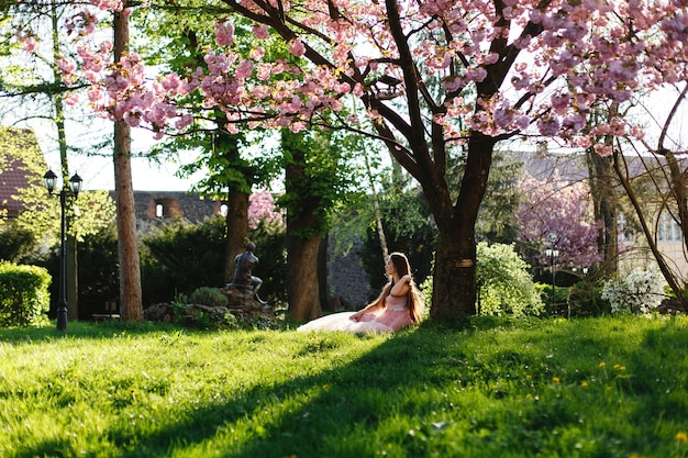 Free photo girl in pink dress sits under the blooming sakura tree in the park