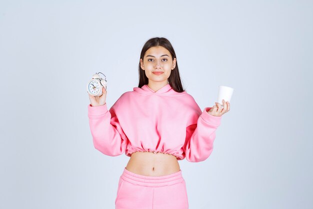 Girl in pink pajamas holding an alarm clock and a cup of coffee. 