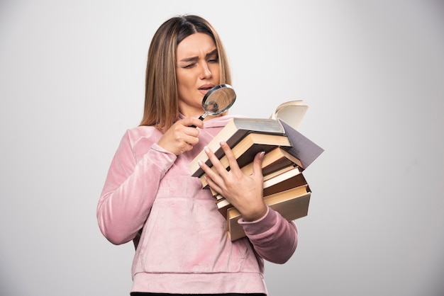 Free Photo girl in pink swaetshirt holding a stock of books and trying to read the top one with a magnifier.