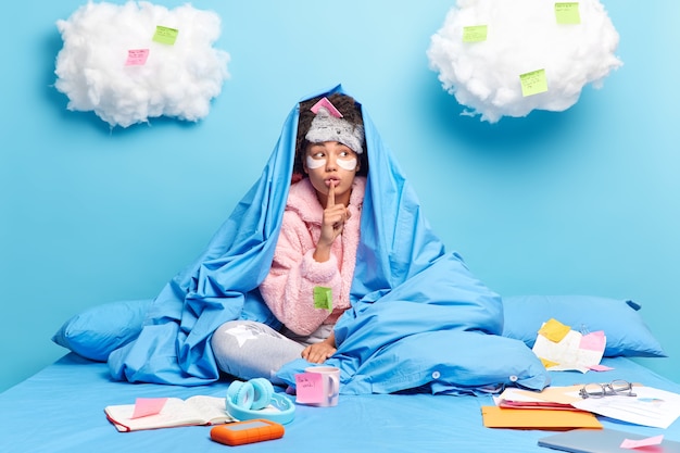 Free photo girl poses on comfortable bed at home studies distantly surrounded by many papers and sticky notes makes secret gesture isolated on blue