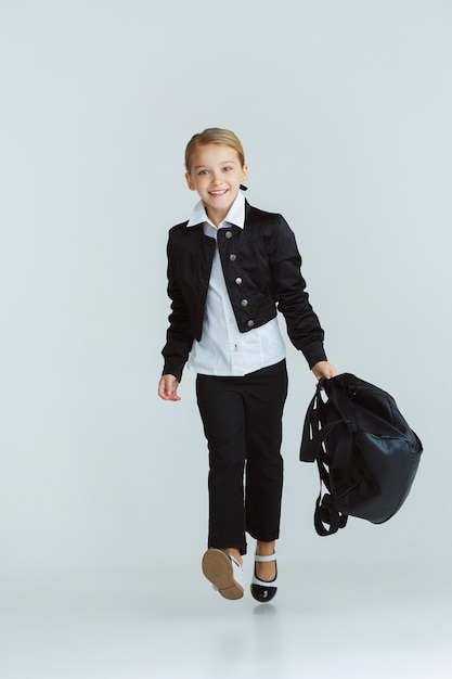 Free Photo girl preparing for school after a long summer break. back to school. little female caucasian model posing in school's uniform with backpack on white wall. childhood, education, holidays concept.