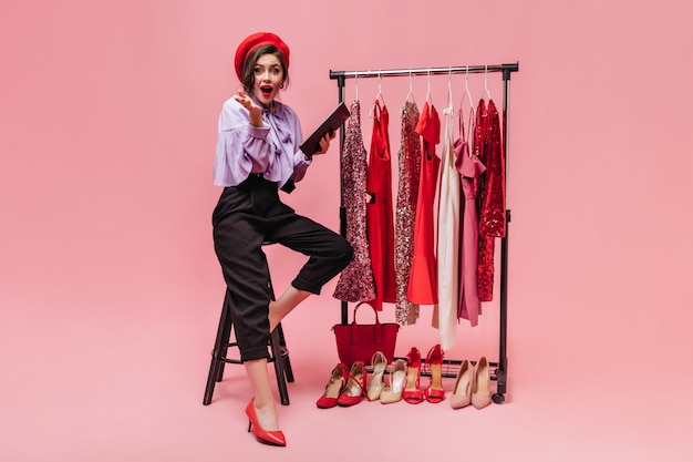 Girl in purple blouse and black pants is sitting on chair, looking into camera and holding folder against background of hangers with bright dresses.