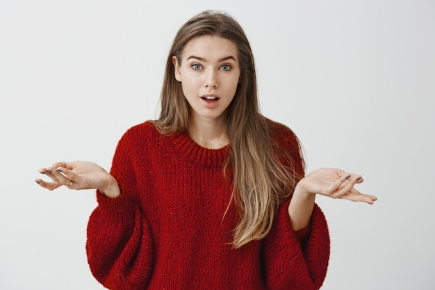 Free photo girl retelling amazing story that happened with her in mall. excited good-looking female student in red loose sweater, shrugging with spread palms, wondering and feeling amazed over gray wall