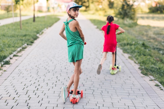 Girl riding on push scooter with her friend on walkway in the park