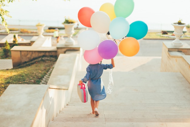 Free photo girl running on the stairs holding colorful balloons and childish suitcase