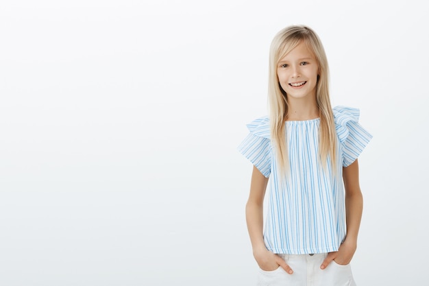 Free Photo girl saved cat from tree, feeling proud. cute little blond kid in stylish blouse, holding hands in pockets and smiling broadly, talking with favorite teacher about good marks, standing over gray wall