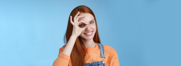 Free photo girl search perfection charming pretty glad redhead female student assuring everything ok showing okay sign eye look through smiling delighted express positive attitude blue background