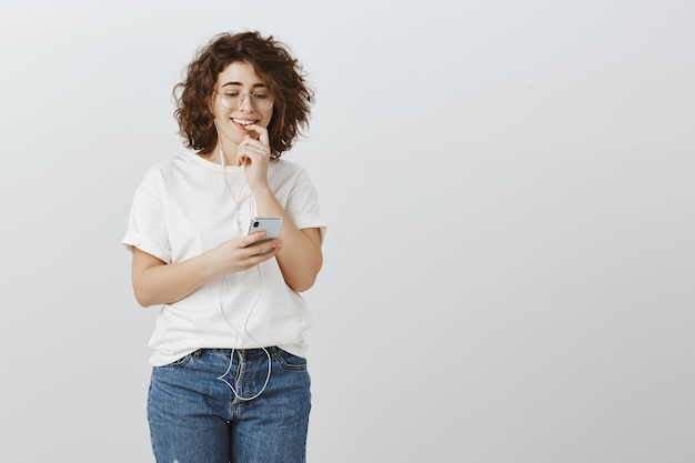 Girl sending risky text, looking excited at smartphone, wearing earphones