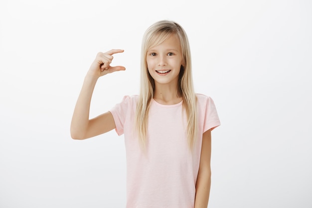 Free photo girl showing how much effort costs to be happy. indoor shot of bright friendly blond female child in pink t-shirt, shapink something small or little and smiling broadly, being excited and joyful