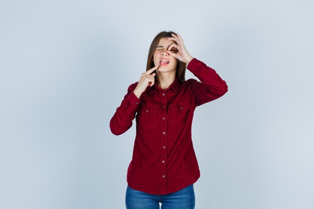 girl showing ok sign on eye, pointing at her tongue in casual shirt and looking pensive 