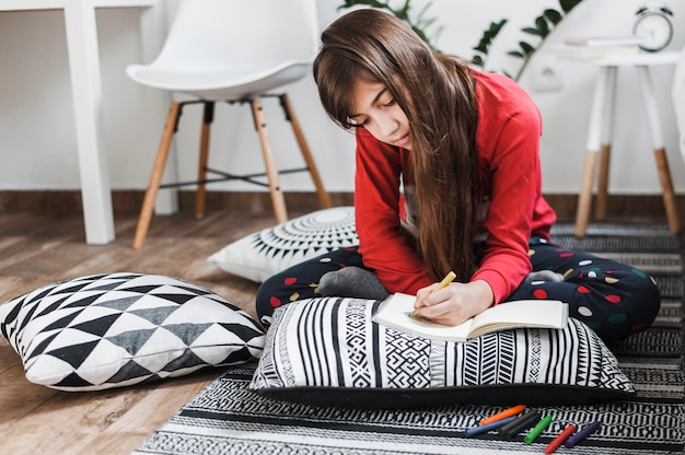 Free photo a girl sitting on rug drawing with colorful crayons on notebook