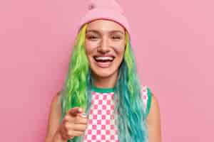 Free photo girl smiles broadly points at you asks to join her team has bright colored hair piercing in nose wears hat checkered shirt stands on pink