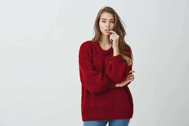 Girl in store tries to pick new shoes. Studio shot of good-looking urban girl in red loose sweater standing with crossed arm and hand on lip, choosing from variants while looking focused 