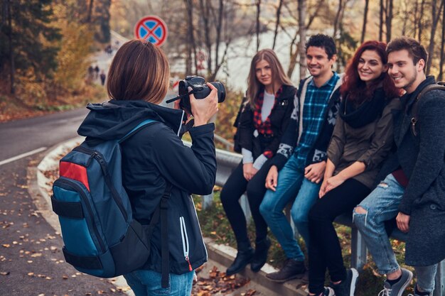 Girl taking a photo of her friends. Group of young friends sitting on guardrail near road. Travel, hiking, adventure concept.