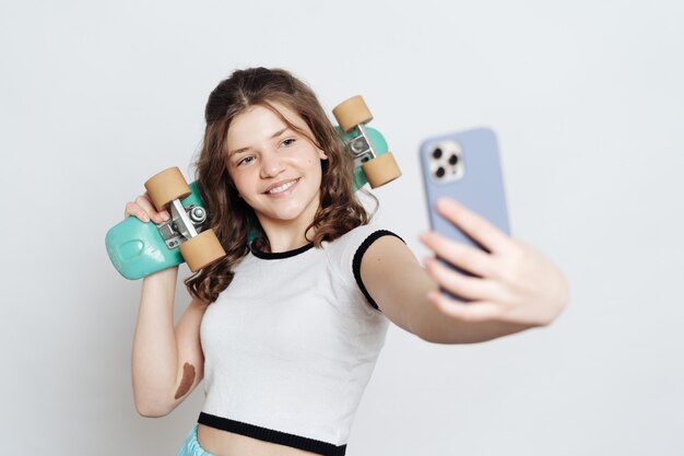 Girl teenager taking selfie on phone while posing with blue penny board on white