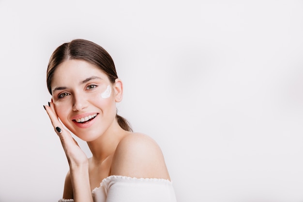 Girl touches moisturized skin and smiles. Portrait of model with cream on face on isolated wall.