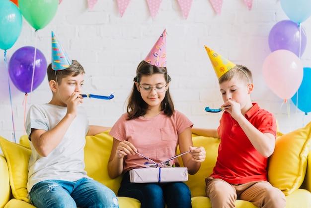 Girl unwrapping birthday gift while her friends blowing party horn