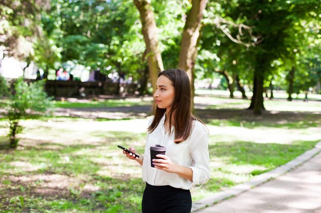 Girl walks with phone in her hand and a cup of coffee in the park