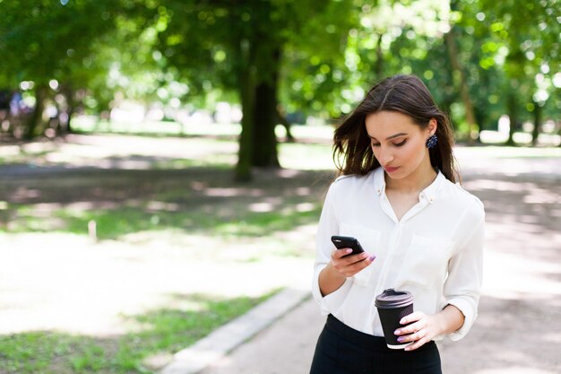 Girl walks with phone in her hand and a cup of coffee in the park