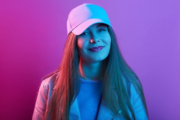 Girl wearing cap and leather jacket looking at camera with charming smile, has pink pomade, standing against pink neon