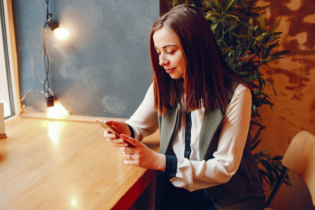 girl in a white blouse sitting at the table near the window in the cafe and holds the phone