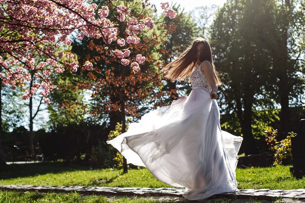 Free photo girl in white dress stands under pink blooming sakura tree in the park