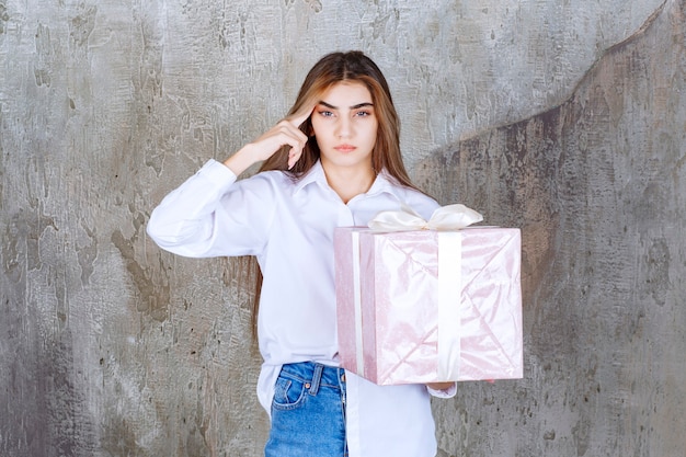 Girl in white shirt holding a pink gift box wrapped with white ribbon and looks confused and hesitating.